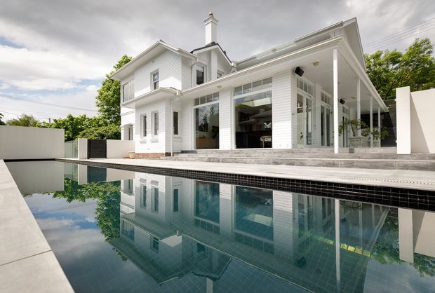 Exterior view of a large white two-storey residence beside a reflective swimming pool. The home features white horizontal weatherboard cladding, a prominent chimney, pitched roof sections, extensive glazed openings, and a covered veranda or terrace supported by slender white columns. Grey paving surrounds the pool, with landscaped greenery and an overcast sky in the background.