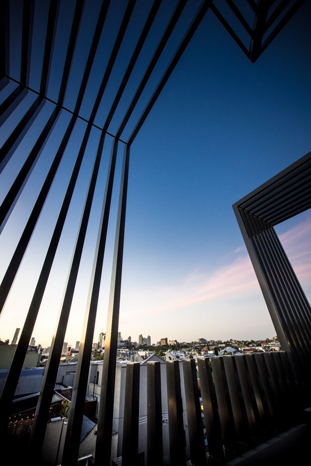 A dramatic architectural view from an elevated balcony or terrace, framed by strong dark vertical and angled metal fins. The open structure creates a geometric portal against a deep blue evening sky, with a dense suburban and urban skyline visible beyond. The image has a high-contrast, contemporary architectural character.