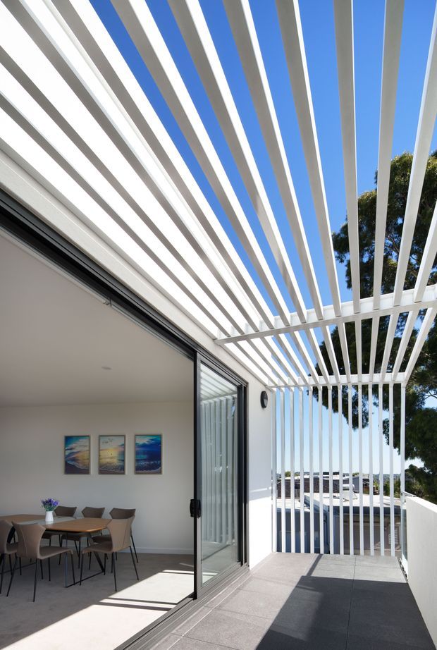 Bright architectural photograph of a contemporary outdoor terrace or balcony adjoining a dining room through large black-framed sliding glass doors. A white open pergola with closely spaced linear rafters creates strong striped shadows across the grey tiled terrace. The interior contains a dining table and chairs, with white walls and framed artwork; trees and blue sky are visible beyond the terrace.