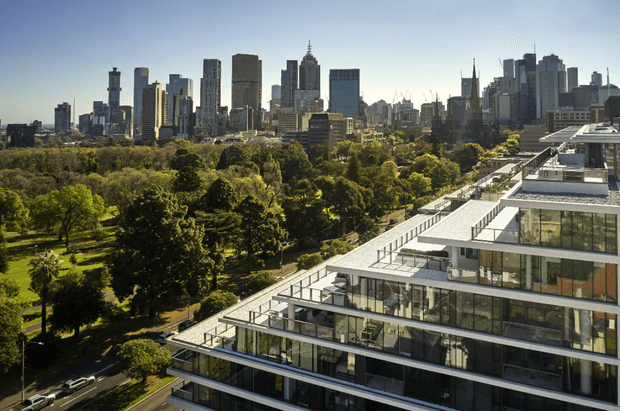 Aerial view of a contemporary multi-storey building with extensive floor-to-ceiling glazing, stacked projecting terraces, white horizontal slab edges and rooftop outdoor areas. A large leafy urban park and dense city skyline are visible beyond.