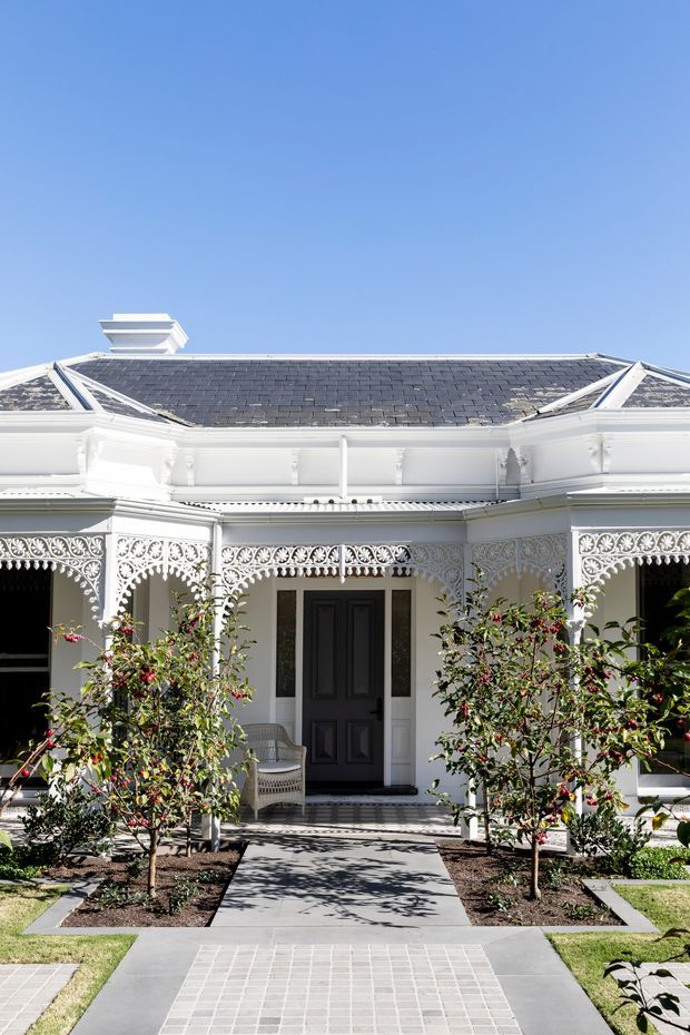 Front exterior view of a restored single-storey residence with ornate white decorative cast-iron verandah detailing, a dark slate hipped roof, white facade, and a central black entrance door. A formal paved walkway leads through landscaped garden beds to the covered porch.