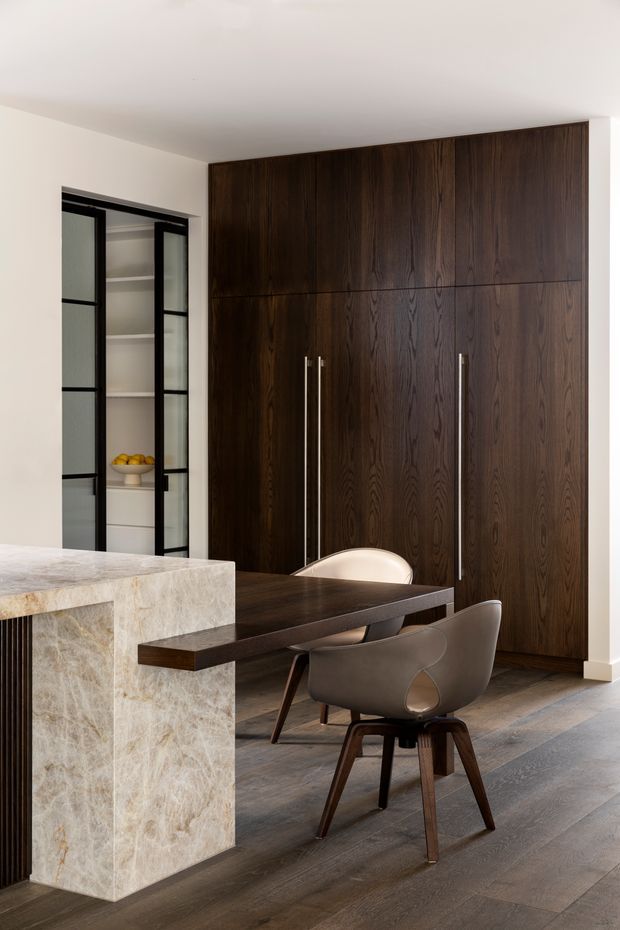 A refined open-plan kitchen and dining area featuring a large pale natural-stone island, a dark timber dining table, sculptural upholstered dining chairs, and a full-height wall of dark timber cabinetry with long vertical handles. Black-framed glazed pantry or storage doors open onto white shelving. Wide-plank timber flooring and warm neutral walls create a sophisticated, warm contemporary interior.
