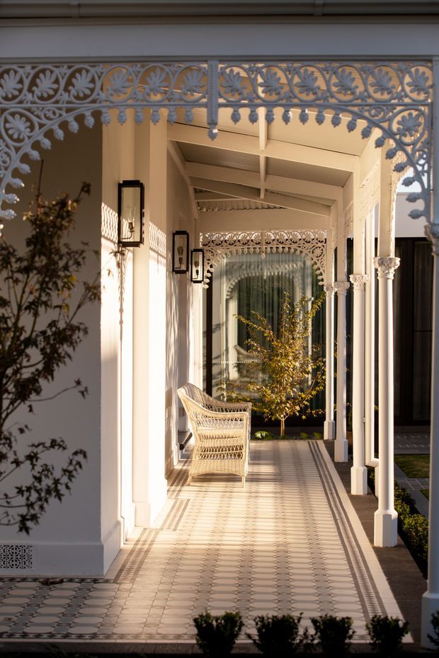 A long covered exterior veranda or walkway with ornate perforated metal arches, slender white columns, decorative capitals, exposed painted roof rafters, wall-mounted lanterns, and patterned geometric tile flooring. A woven outdoor chair sits along the passage, which opens toward a landscaped courtyard and glazed doorway. The architecture has strong heritage-inspired decorative detailing and a warm, elegant atmosphere.