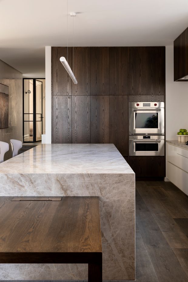 A contemporary kitchen dominated by a large pale veined stone island with an integrated dark timber dining table extension. Full-height dark wood cabinetry forms the rear wall and incorporates stacked stainless-steel ovens. Pale cabinetry, dark timber flooring, a slim linear pendant, and black-framed glazed doors create a refined, warm-toned interior.