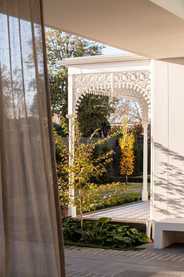 A refined view from an interior through a covered opening toward a landscaped courtyard. A sheer curtain frames the left side, while a white rendered wall and ceiling surround an ornate white decorative metal arch or gate with intricate scrollwork. The courtyard contains structured garden beds, leafy planting, and patterned paving, with warm late-afternoon light creating a calm, elegant atmosphere.