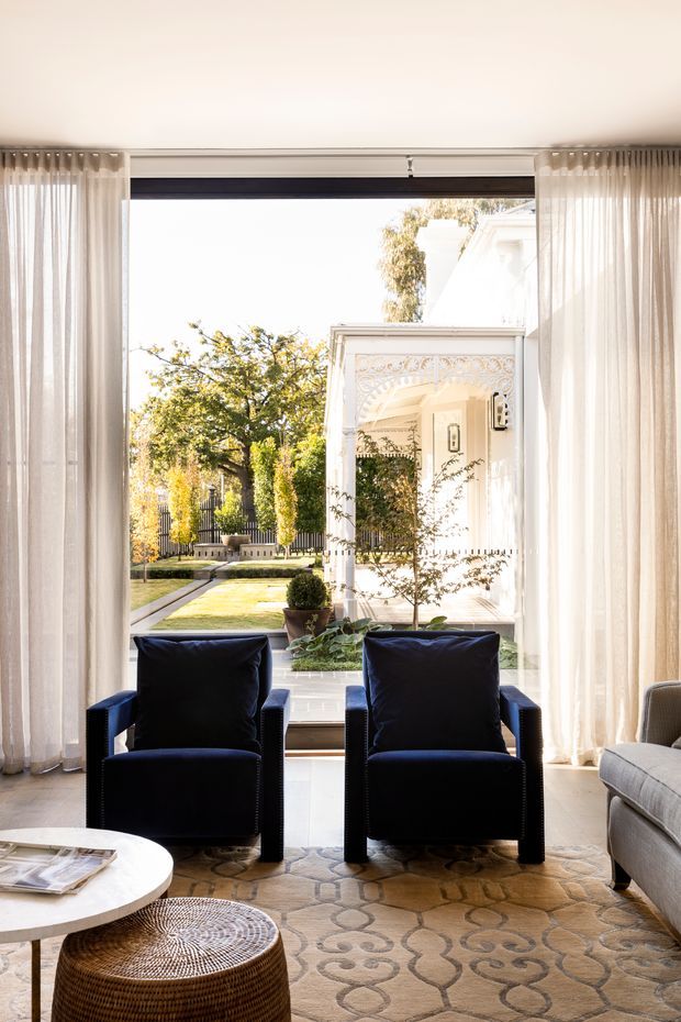 Bright, refined living room with two dark upholstered armchairs positioned in front of full-height glazing and sheer curtains. A pale sofa, round white coffee table, woven ottoman, and patterned neutral rug furnish the room. The glazing overlooks a landscaped garden and a highly ornate white architectural facade with decorative moulding and columns.