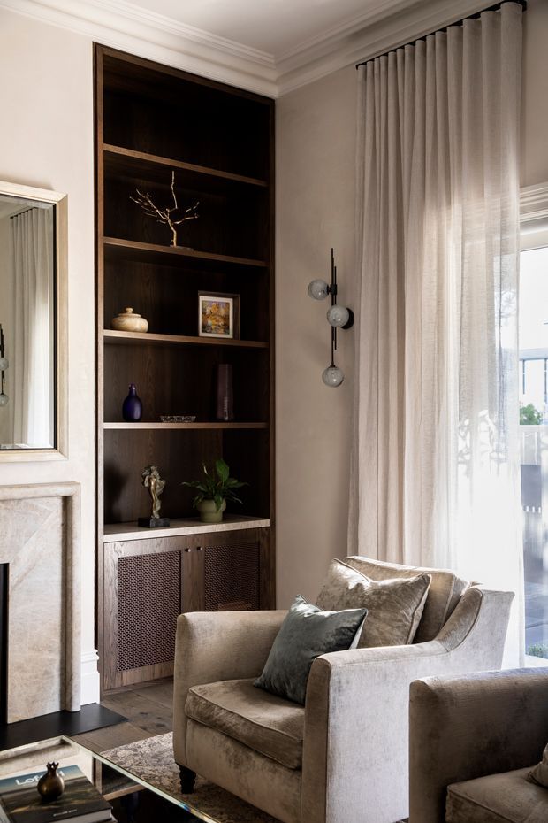 A refined living room corner featuring a deep dark-wood built-in shelving and cabinetry unit with decorative objects, a pale stone fireplace surround at left, a large framed mirror, and a plush neutral upholstered armchair. Floor-to-ceiling sheer curtains soften the bright window, while a sculptural dark metal wall light and glass coffee table add contemporary detailing. The room has traditional architectural trim combined with elegant contemporary furnishings and warm layered neutral tones.