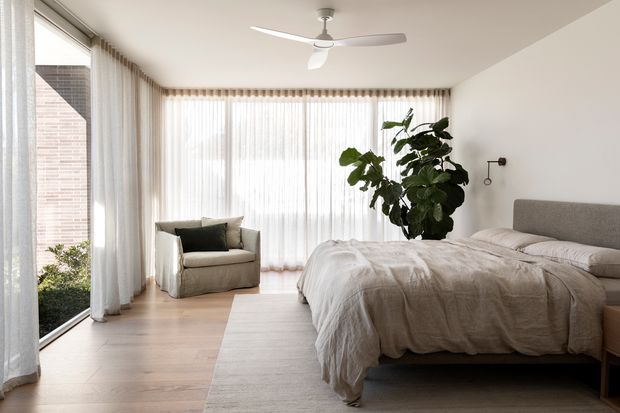Bright, airy contemporary bedroom with a large upholstered bed, layered neutral bedding, a sculptural armchair, sheer floor-to-ceiling curtains, pale timber flooring, and abundant natural light. A large potted fiddle-leaf fig and minimalist wall light add restrained organic detail. The room opens visually to an exterior brick wall and landscaping through expansive glazing.