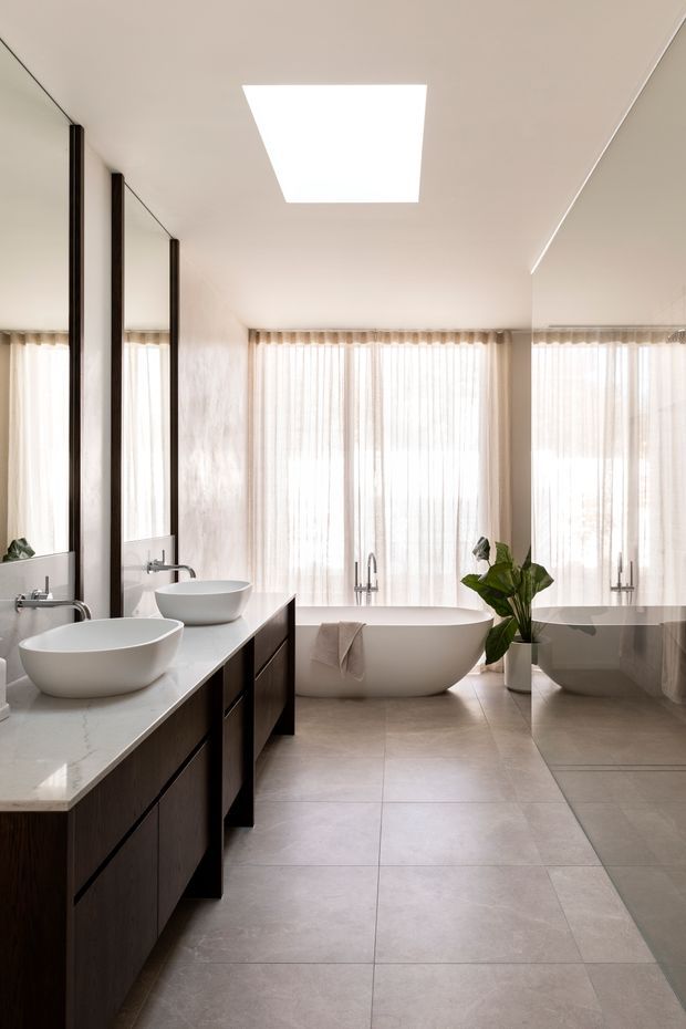 A bright, refined contemporary bathroom with a freestanding white bathtub positioned in front of sheer full-height windows. A long dark timber vanity with two white vessel basins and wall-mounted chrome taps runs along the left wall, while large mirrors reflect the room. The space has pale large-format tiled flooring, a smooth light ceiling with a rectangular skylight or ceiling light, soft natural light, and a potted plant near the tub.