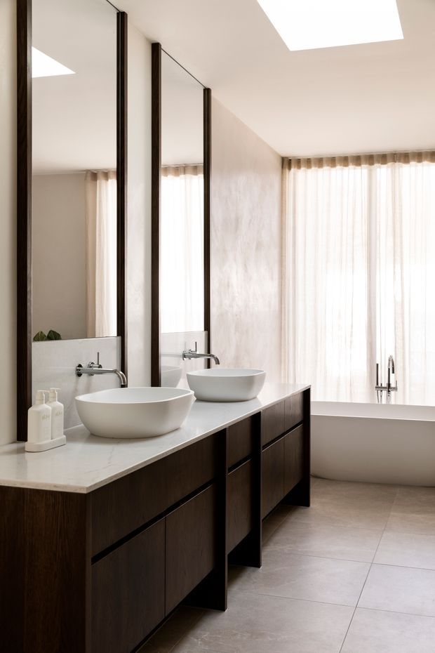 A bright, refined bathroom featuring a long double vanity with two white vessel basins, dark timber cabinetry, a pale stone benchtop, wall-to-wall mirrors, and brushed metal tapware. Large sheer-curtained windows and a freestanding bathtub are visible at the rear, with a rectangular skylight providing additional daylight. The room has large pale floor tiles and a warm neutral palette.