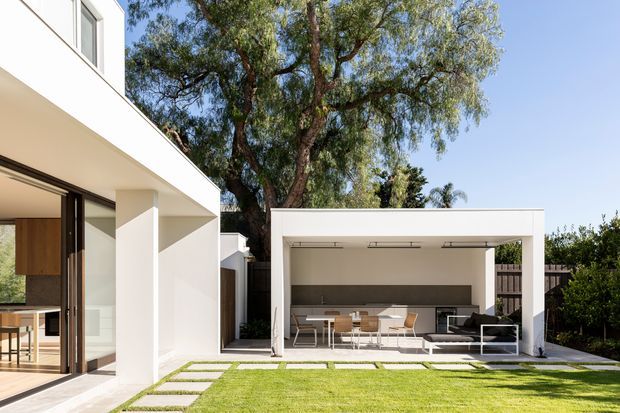 Bright contemporary residential exterior showing a white two-storey house opening onto a landscaped lawn and a separate covered outdoor entertaining pavilion. The pavilion contains a dining table, chairs, a long built-in counter or barbecue zone, and outdoor seating. Large sliding glass doors connect the main house to the terrace, while stepping stones cross the grass. Mature trees and boundary planting frame the scene.