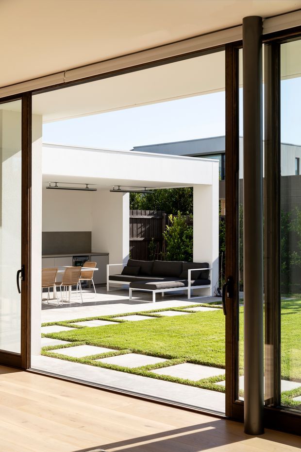 Bright contemporary indoor-outdoor living space viewed from inside through expansive dark timber-framed glazing. A light timber floor opens onto a manicured lawn with large square stepping stones, leading to a covered outdoor entertaining area with a built-in kitchen, dining setting, and black outdoor sofa. Clean white walls, flat rooflines, strong horizontal and vertical lines, and abundant natural light create a refined connection between the interior and courtyard.