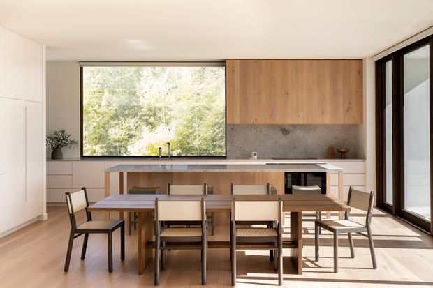 Bright, open-plan kitchen and dining area with a long central island, integrated cabinetry, timber feature cabinetry, stone-look splashback, and a dining table surrounded by simple timber and woven-seat chairs. Large black-framed windows and glazed doors provide views to greenery and abundant natural light. The space has a clean, restrained contemporary character with warm timber finishes.