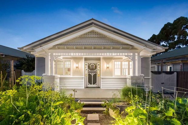 Front exterior view of a single-storey heritage-style cottage or bungalow, featuring a symmetrical gabled verandah, white weatherboard cladding, decorative timber fretwork, columns, panelled entry door, and illuminated garden beds leading to the front steps.