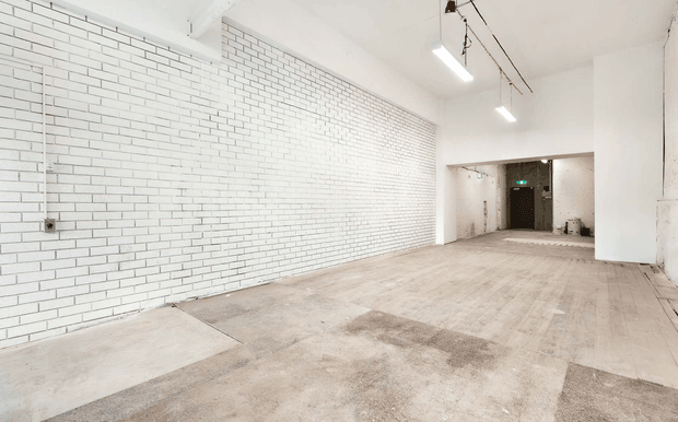 Empty, elongated commercial or industrial interior with a white-painted brick feature wall, exposed concrete floor, white walls, and simple suspended linear lighting. A recessed doorway and service area are visible at the far end, creating a spacious gallery, studio, or warehouse-like setting.
