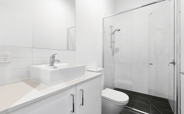 Bright, compact contemporary bathroom with a white vessel basin set on a white vanity, mirrored wall cabinet, wall-hung toilet, and frameless glass shower enclosure. White tiled walls and cabinetry contrast with dark floor tiles and chrome fittings.