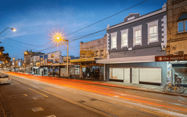 Evening streetscape of a dense urban commercial strip, with historic two-storey shopfront buildings, illuminated retail signage, overhead utility wires, street lighting, and long-exposure vehicle light trails along the roadway.