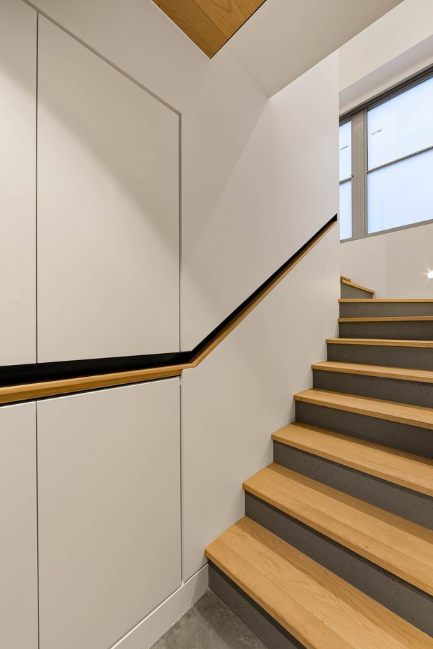 Contemporary interior stairway with a clean, minimalist fit-out. The staircase combines warm timber treads and handrail detailing with dark grey risers, white integrated wall cabinetry, and a large window bringing in natural light. A timber-lined ceiling inset adds a subtle architectural feature.