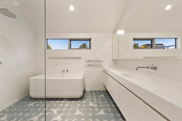 Bright contemporary bathroom with a freestanding white bathtub, double vanity with integrated basins, wall-mounted fixtures, large mirrored cabinetry, narrow horizontal windows, white tiled walls, and patterned circular floor tiles.