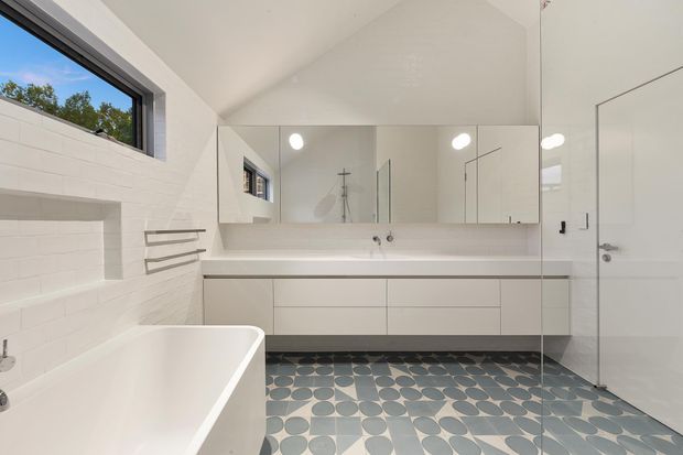 Bright contemporary bathroom with a freestanding white bathtub, large wall-to-wall mirrored vanity, pale built-in cabinetry, white tiled walls, and a distinctive geometric blue-grey patterned floor. A walk-in shower and white door are visible, with natural light entering through a high horizontal window.