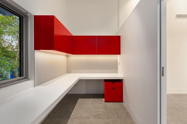 A compact modern workspace with an L-shaped white built-in desk beneath vivid red overhead and drawer cabinetry. Integrated under-cabinet lighting illuminates the pale work surfaces, while a large grey-framed window provides natural light and views of greenery. The room has smooth light walls, a dark grey floor section, and a minimalist built-in design.