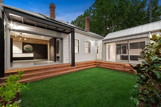 A renovated single-storey residence arranged around a landscaped courtyard. The white weatherboard-clad main building has a pitched metal roof and brick chimneys, while a dark-framed glazed opening connects the interior to a timber deck. A contemporary pavilion with a standing-seam metal roof and large windows forms the opposite side of the courtyard. The visible interior has dark cabinetry or wall panels, pale flooring, and modern lighting.