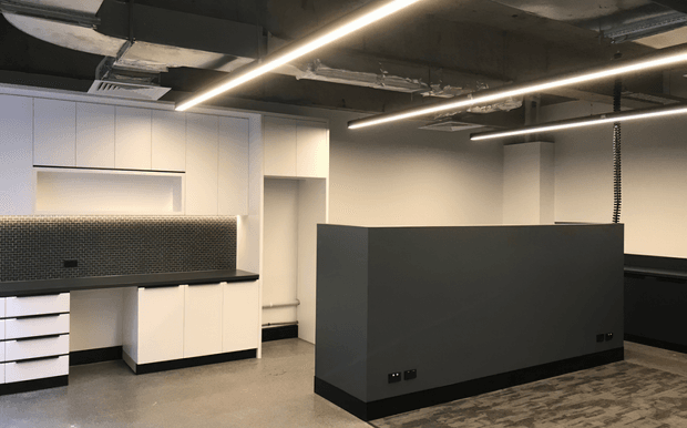 A modern open-plan commercial workspace or office fit-out with white built-in cabinetry, a dark mosaic-tile splashback, black benchtops, a large dark grey central partition or workstation enclosure, exposed concrete services, linear LED lighting, and polished concrete flooring. The palette is monochromatic and industrial, with clearly defined work zones.