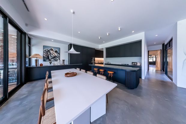 Contemporary open-plan kitchen and dining area with a long white dining table, woven timber dining chairs, dark built-in cabinetry, a large kitchen island with bar stools, pendant lighting, concrete-look flooring, and extensive black-framed glazing. A hallway and additional interior spaces extend beyond the kitchen.