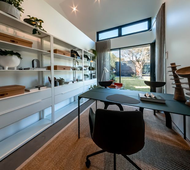 A bright, contemporary dining and living space with a dark dining table and upholstered chairs, extensive white built-in shelving and cabinetry, decorative objects and plants, and full-height glazing opening to a landscaped garden. High clerestory windows and layered curtains bring in abundant natural light.