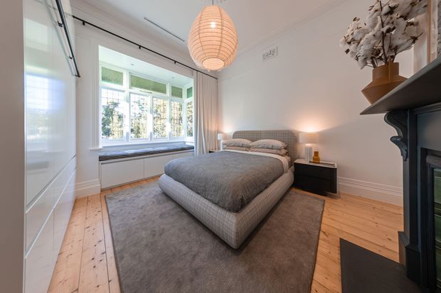 A refined bedroom with a large upholstered bed centered on a neutral rug, pale timber floorboards, white walls, and a bright bay window with built-in window seating or cabinetry. A black fireplace surround and dark shelving create contrast, while a woven pendant light and soft bedside lighting add warmth. The room combines classic architectural detailing with contemporary furnishings.