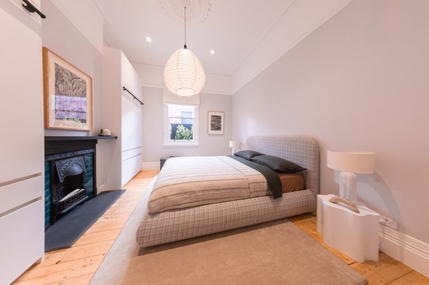 Bright, refined bedroom with a large upholstered plaid bed, neutral bedding, timber floors, a traditional dark fireplace, white built-in cabinetry, and soft contemporary lighting. The room combines classic architectural details with understated contemporary furnishings.