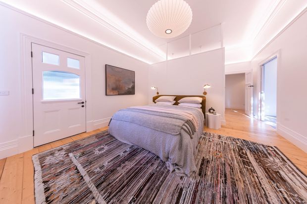Bright, refined bedroom with a centrally placed upholstered bed, layered neutral bedding, a large abstract patterned rug, pale timber flooring, white paneled walls and doors, decorative ceiling mouldings, and a sculptural pendant light. A hallway and adjoining doorway are visible in the background.