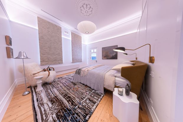 A refined bedroom with a warm, layered palette and contemporary detailing. The room features a timber floor, pale walls with decorative ceiling mouldings and a central ceiling medallion, textured wall panels behind the bed and on the opposite wall, a sculptural pendant light, a large abstract-patterned rug, and a bed with an upholstered mustard-brown headboard. Minimal bedside furniture, a floor lamp, framed artwork, and soft neutral textiles complete the space.