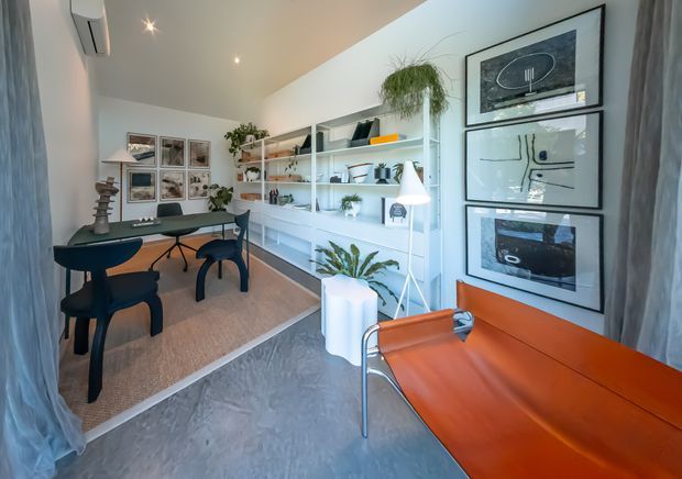 Bright, compact contemporary home office with a long white built-in shelving and desk wall, framed artwork, plants, task lighting, and a small meeting or dining table with dark chairs. The room has polished grey flooring, a warm neutral rug, sheer curtains, and a vivid orange leather bench or sofa in the foreground.