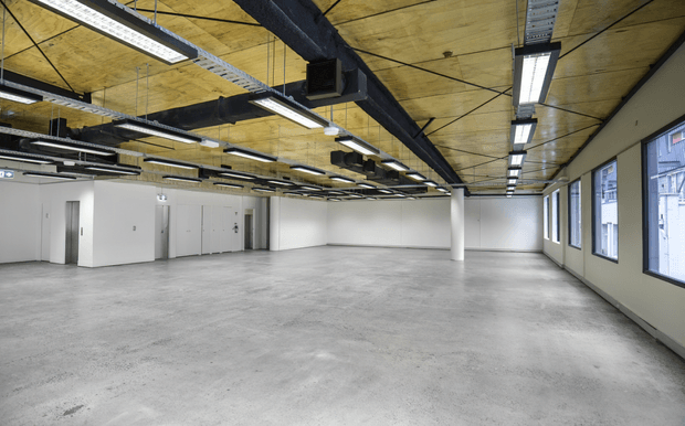 Large open-plan commercial interior with a raw concrete floor, plain white walls, exposed dark mechanical services and ductwork, and a distinctive wood-panelled ceiling grid. Long linear fluorescent and LED fixtures provide bright, even illumination, while large windows line one side of the space. The room appears vacant and suitable for office, studio, or warehouse-style use.