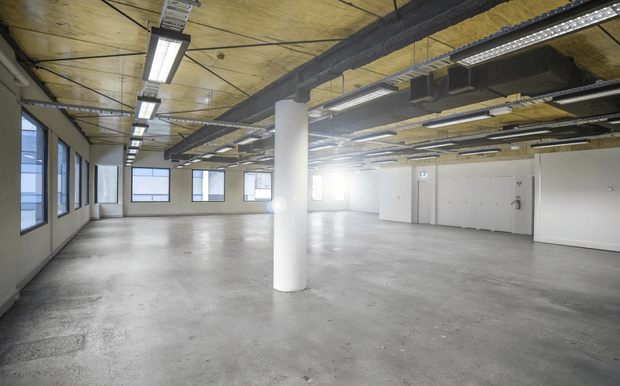 A large open-plan commercial interior with a raw concrete floor, exposed dark structural beams and services, a plywood-toned ceiling, fluorescent strip lighting, white cylindrical columns, extensive windows, and enclosed white utility or office rooms along one side. The space appears vacant and adaptable, with an industrial warehouse or office fit-out character.