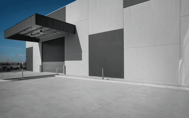Exterior view of a large modern industrial or commercial building with a minimalist white and dark-grey panelled facade. The building features a projecting loading-canopy structure, a roller shutter door, exposed service fixtures, broad concrete paving, and clean geometric lines. The image is predominantly monochrome with a small area of blue sky visible.