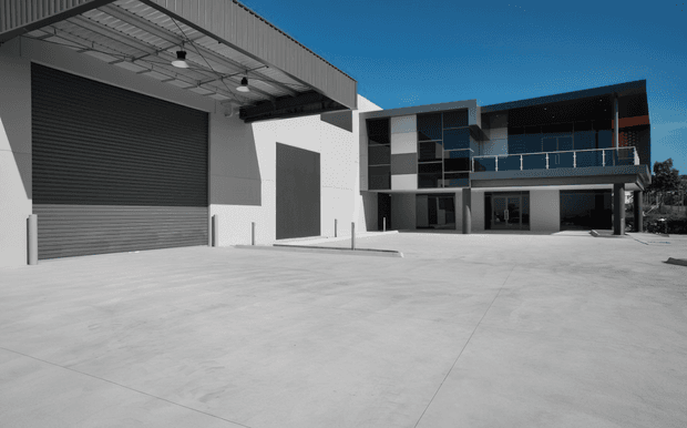 A modern commercial or industrial building viewed from a broad concrete forecourt. The composition includes a large roller shutter loading-bay door beneath a covered steel canopy, with exposed ceiling services and pendant lights. A two-storey office or workshop block sits behind, featuring dark and light rectangular facade panels, extensive glazing, an upper balcony with glass balustrade, and an open ground-level undercroft.
