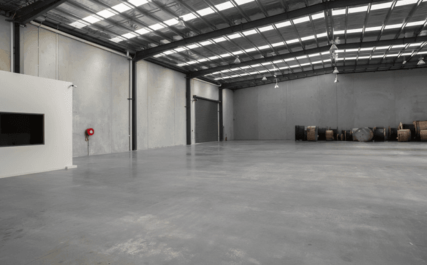 A large, mostly empty industrial warehouse interior with a polished concrete floor, exposed steel roof structure, translucent roof panels, concrete walls, roller doors, hanging industrial lights, and a small white partition containing a wall-mounted screen. A row of industrial drums or equipment sits along the rear wall.