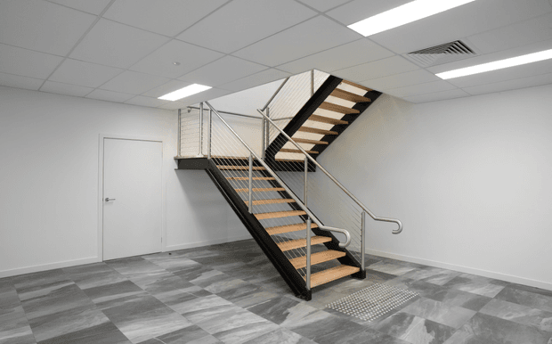 An empty commercial interior featuring a modern U-shaped metal staircase with timber stair treads and cable railings. The space has white walls, a suspended acoustic tile ceiling with fluorescent lighting, a white door, and grey patterned carpet tiles. A tactile floor indicator is positioned at the base of the stairs.