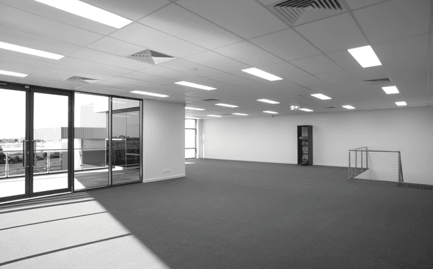 A large, empty commercial office interior with grey carpet, white walls, suspended acoustic ceiling tiles, recessed fluorescent lighting, exposed HVAC grilles, and extensive black-framed glazed doors and windows. A stair opening with a simple metal balustrade is visible at the right, and strong daylight enters from the glazing.