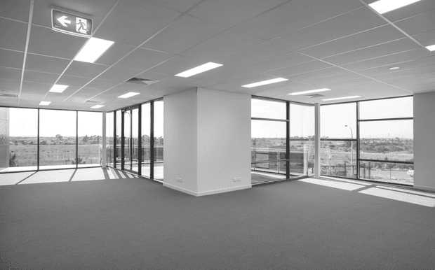 Bright, empty commercial office interior with expansive floor-to-ceiling glazing, carpeted floors, white walls, suspended acoustic ceiling tiles, and recessed fluorescent lighting. The open-plan space has panoramic urban views and a glass-enclosed meeting room or office around a central partition wall. An illuminated emergency exit sign is visible near the ceiling.