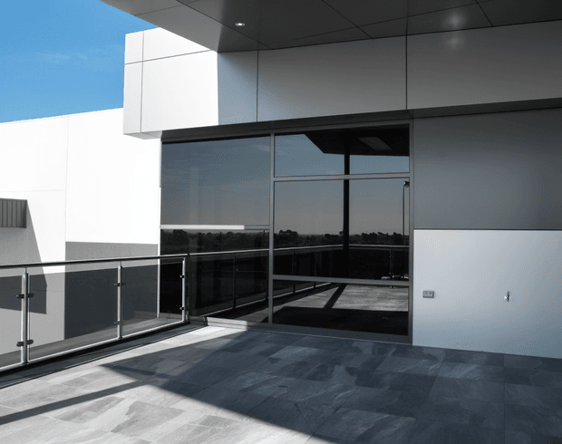 Modern rooftop terrace or balcony with dark slate-like tiled flooring, frameless glass balustrade, large dark-framed glazing, and crisp white and grey contemporary building surfaces. The terrace is brightly lit with strong architectural shadows and expansive reflected views through the glazing.
