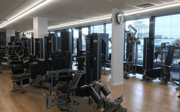 A spacious modern commercial fitness gym filled with rows of weight-training and resistance machines. The room has timber-look flooring, dark exercise equipment, white structural columns, a suspended grid ceiling with linear lighting, large floor-to-ceiling windows, and an illuminated wall clock. The space appears clean, bright, and professionally fitted out.