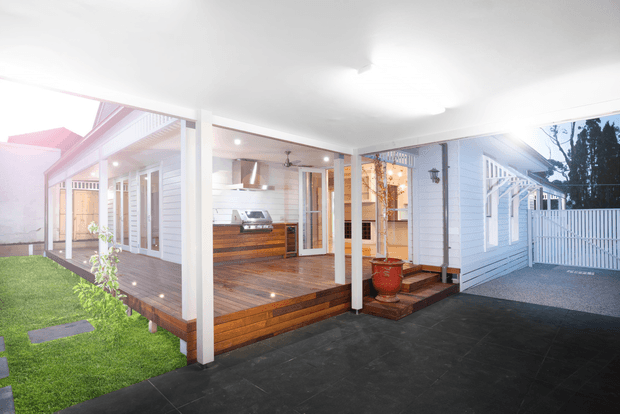 A covered outdoor entertaining area and carport beside a light-coloured weatherboard house. The space features a timber deck, built-in outdoor kitchen with stainless-steel rangehood and cabinetry, steps, white posts and railings, dark paved flooring, and a fenced side yard. Interior kitchen and warm pendant lighting are visible through the glazing.