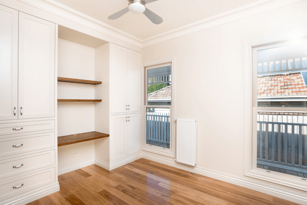 Bright, unfurnished bedroom with built-in white cabinetry and drawers, recessed open shelving with dark timber shelves, polished timber flooring, decorative ceiling cornices, a ceiling fan, and two tall windows overlooking neighbouring rooftops and balconies. A wall-mounted radiator sits between the windows.