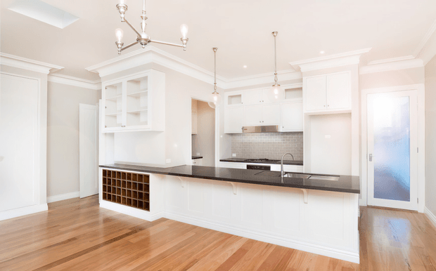Bright, spacious white kitchen with a large island featuring a dark stone benchtop, integrated sink and panelled cabinetry. The room has warm timber flooring, a tiled splashback, glass-fronted display cabinetry, decorative ceiling mouldings, pendant lights, and a frosted-glass door. A built-in wine rack is visible beneath the island-side cabinetry.