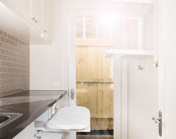 Bright, compact utility or laundry space with white built-in cabinetry, a dark stone benchtop, stainless sink, and grey subway-tile splashback. A white front-loading washing machine is visible beneath the bench, with open shelving and a timber-fenced exterior area beyond the doorway.