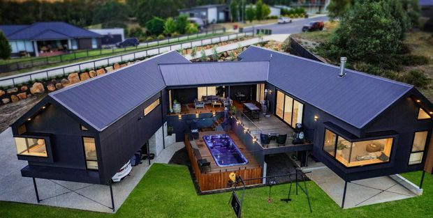Aerial view of a contemporary dark-clad residential home arranged around a central courtyard and outdoor entertaining area. The house has multiple pitched roof wings, extensive glazing, a timber deck with a spa or plunge pool, lawn, and a children's swing set. A suburban hillside neighborhood is visible in the background.