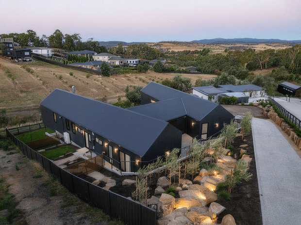 Elevated dusk view of a dark-clad, single-storey residential building with multiple pitched gabled roof forms, warm interior lighting, timber terraces, and a landscaped rock garden along a broad concrete driveway. The home sits on a sloping semi-rural site with scattered neighbouring dwellings and rolling hills in the distance.